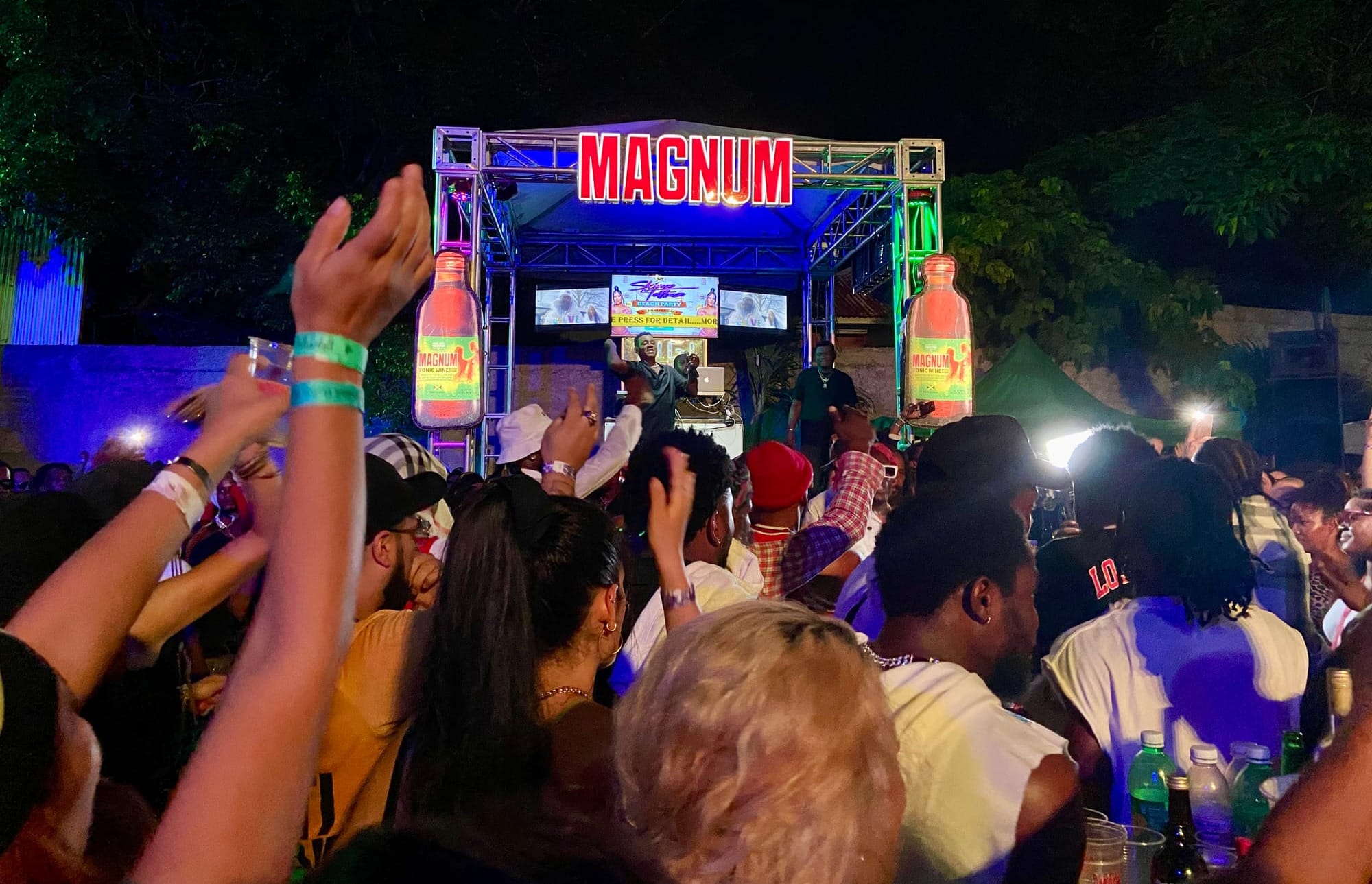 A party outside. People are listening to the music or dancing. There is a stage for the DJ and host. The stage has a "MAGNUM" sign at the top and a magnum bottle sign on the left and right.