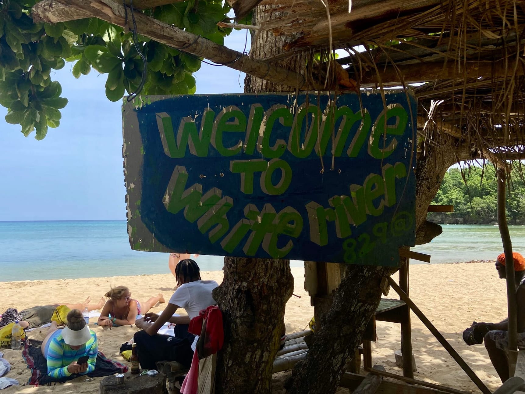 A blue sign with green and white letters on a tree that says "Welcome to White river". In the background is the beach and the sea.