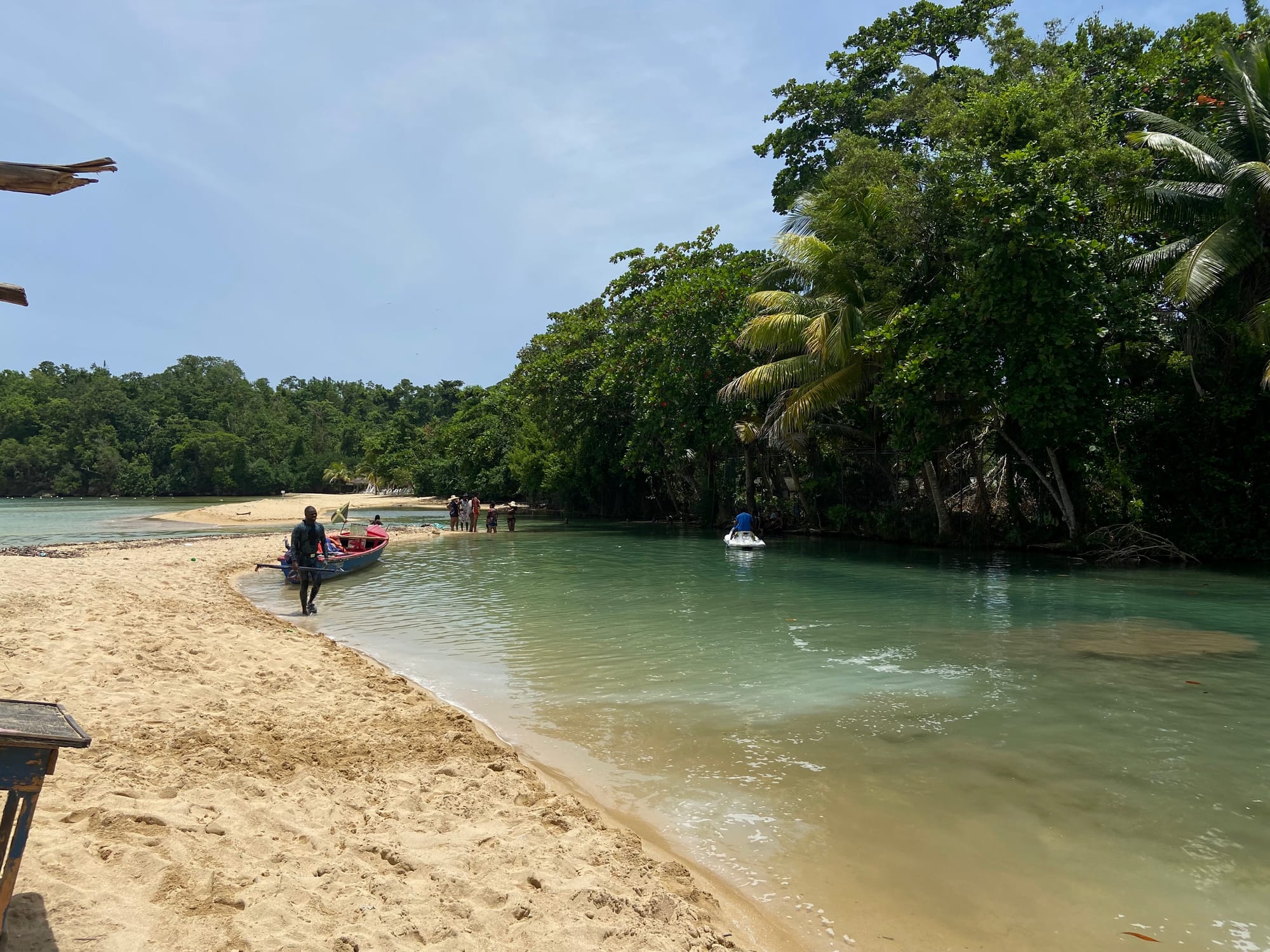 Beach on the left and the river on the right. There are trees on the right side of the river. In the background you see a boat and a jet ski.