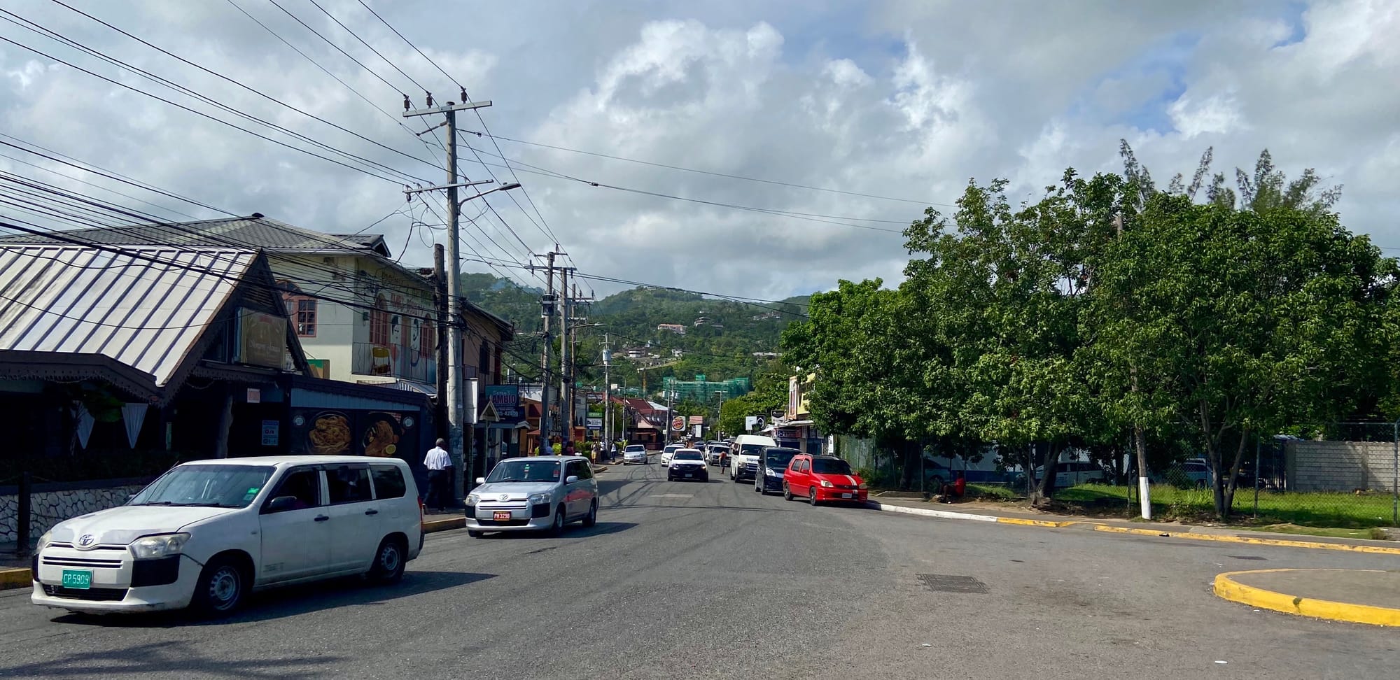 Street with cars 4 cars driving down the road. There are houses on the left and trees on the right. In the back there is the mountainside.