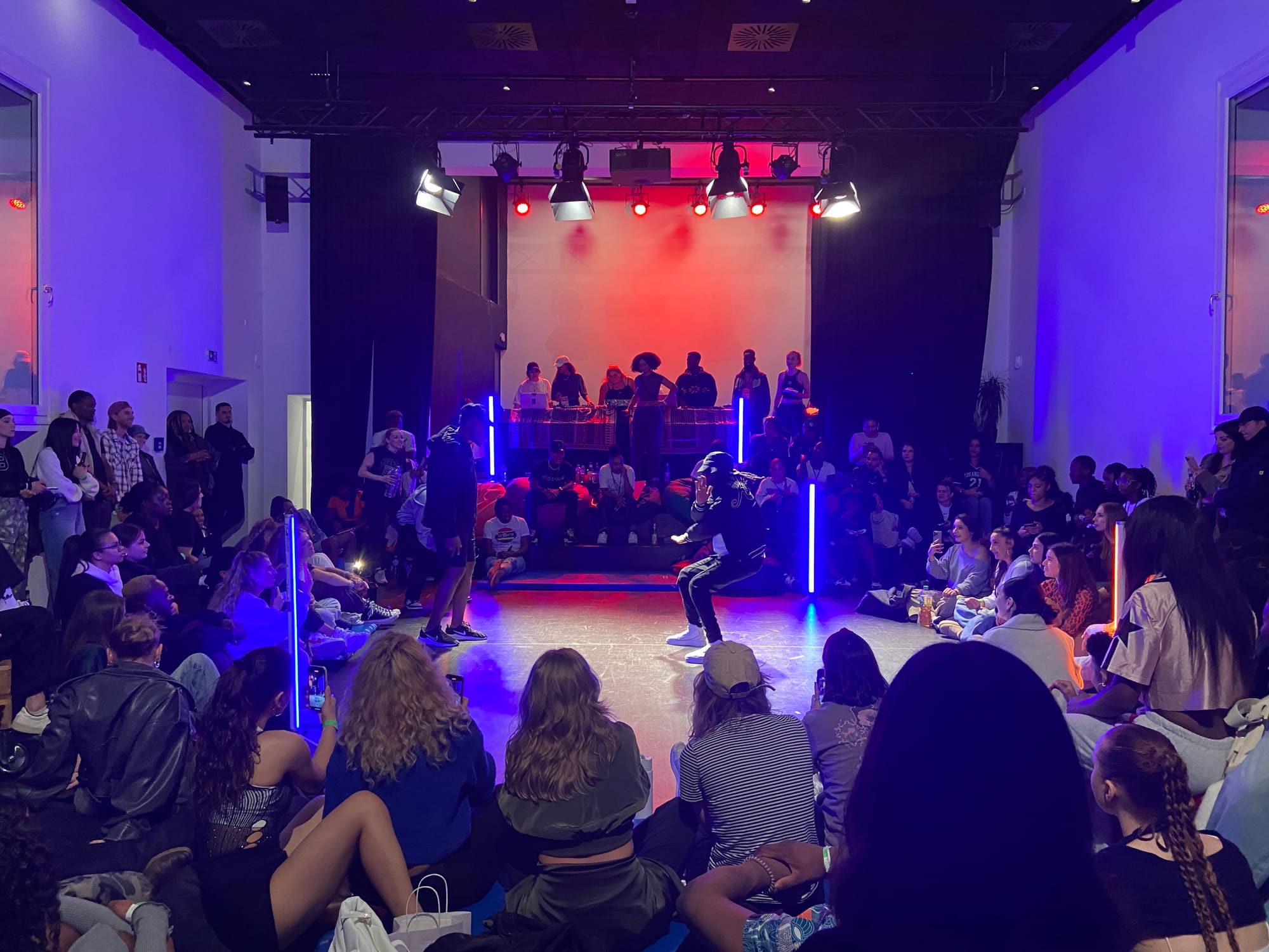 There is a dance floor in the middle. People are seated and standing around the dance floor. Behind the dancers are 3 judges. Behind the judges, on the stage, are the host and the DJ.