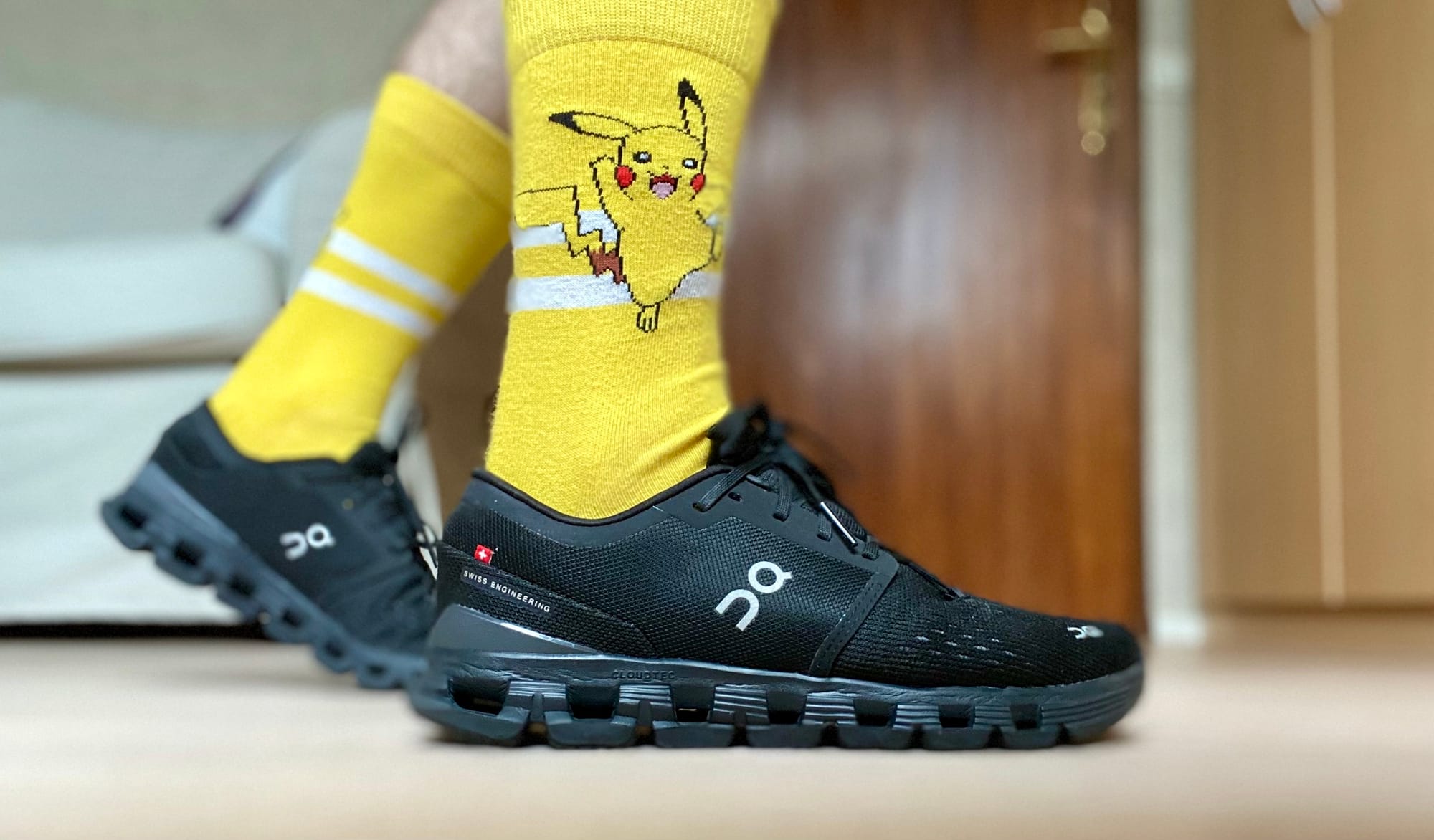 Right shoe is in the front. Left shoe is in the back, heel lifted. The shoes are black with the On logo in with on them. The socks are yellow and have a picture of Pikachu jumping on it.