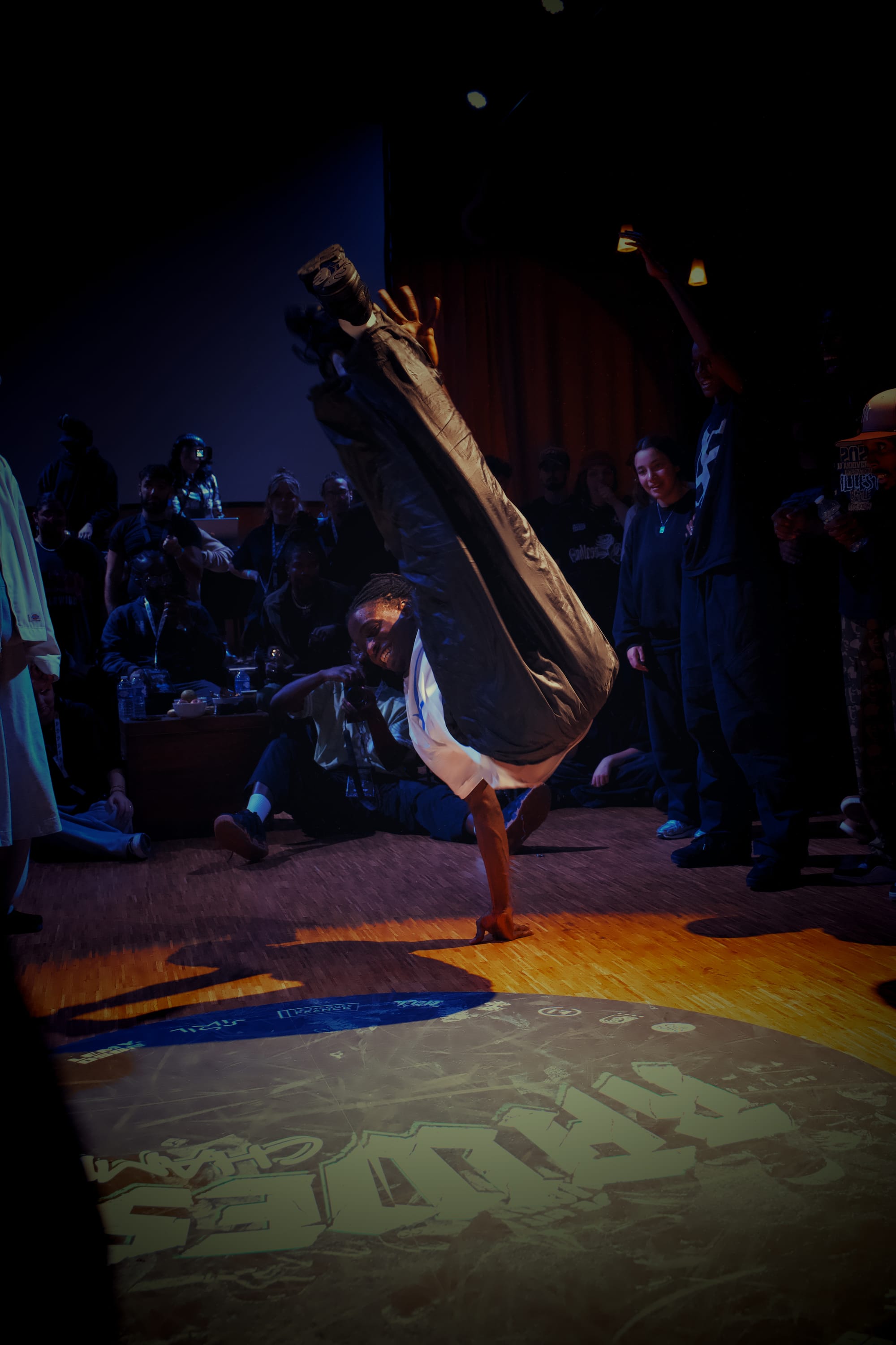 Kimmy Boomclap is dancing during the 6 vs 6 exhibition battle. He is kicking both legs in the air while balancing on his right hand.