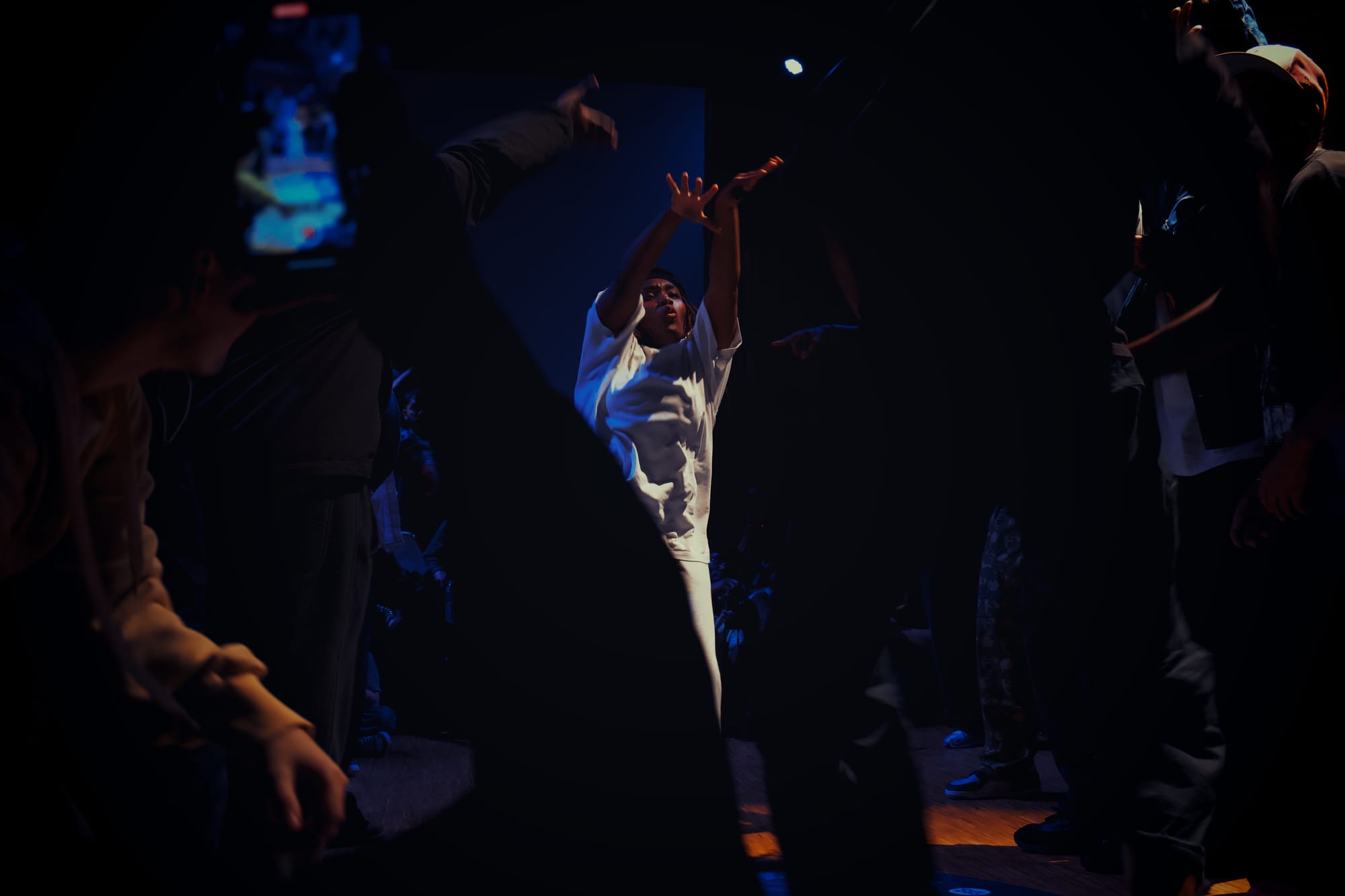 Kassy is dancing during the 6 vs 6 exhibition battle. She is surrounded by the other dancers and the audience.