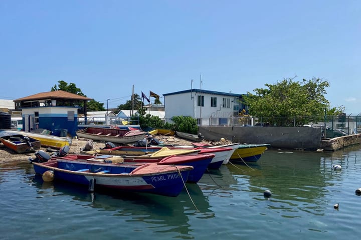 A harbour where you see a part of the sea and part of the shore. There are six boats in the water and couple of baots on the