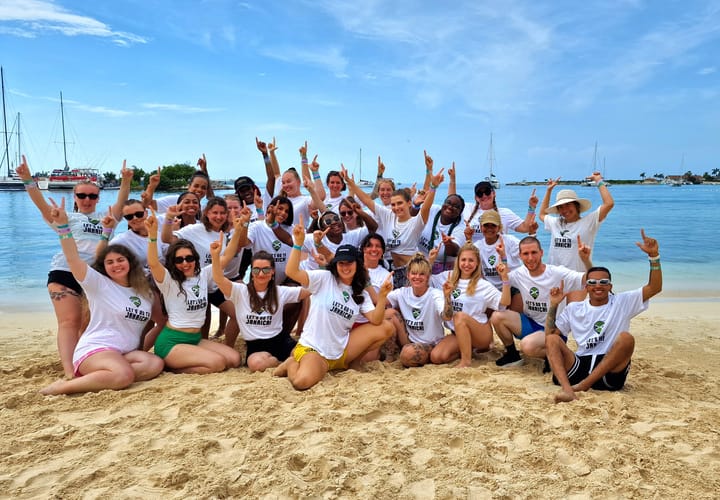 Group picture of 27 people on the beach with their back to the sea. They are pointing both their index fingers up to the sky.