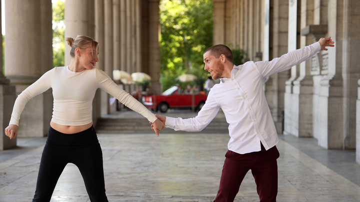 Clara (left) and Slim B (right) dancing. Slim B is holding the hand of Clara, while they are looking at each other.