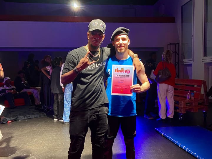 Chin (left) is standing next to Slim B (right). Slim B is holding a certificate proving that he won the Dancehall 1 vs 1.