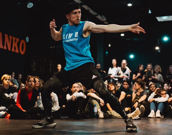 Slim B wearing a blue shirt, black shorts and camo tights is dancing in a battle. There are people seated behind him.